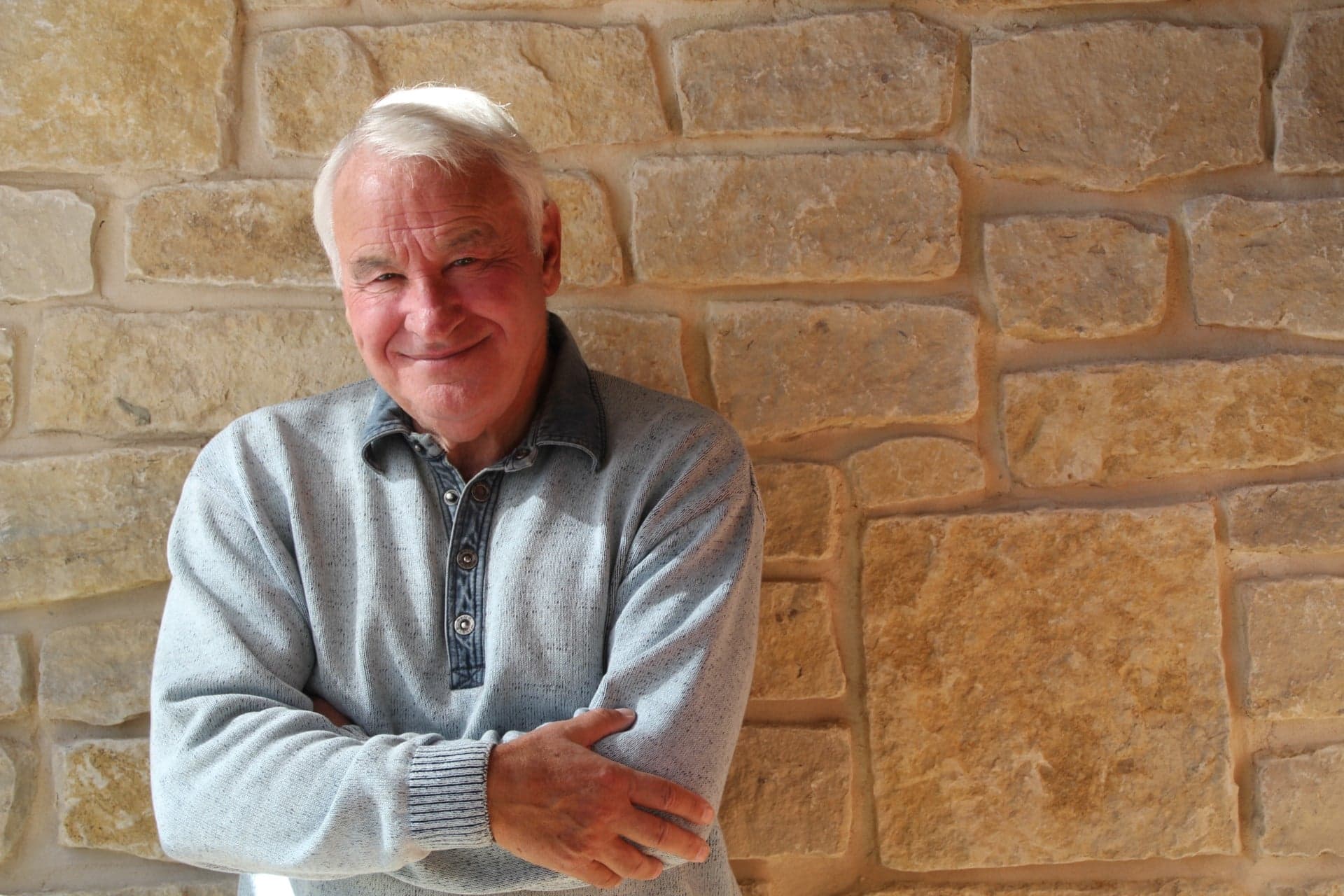 Tom Golisano, a smiling man wearing a gray-collared sweater stands against a stone wall with his arms crossed.